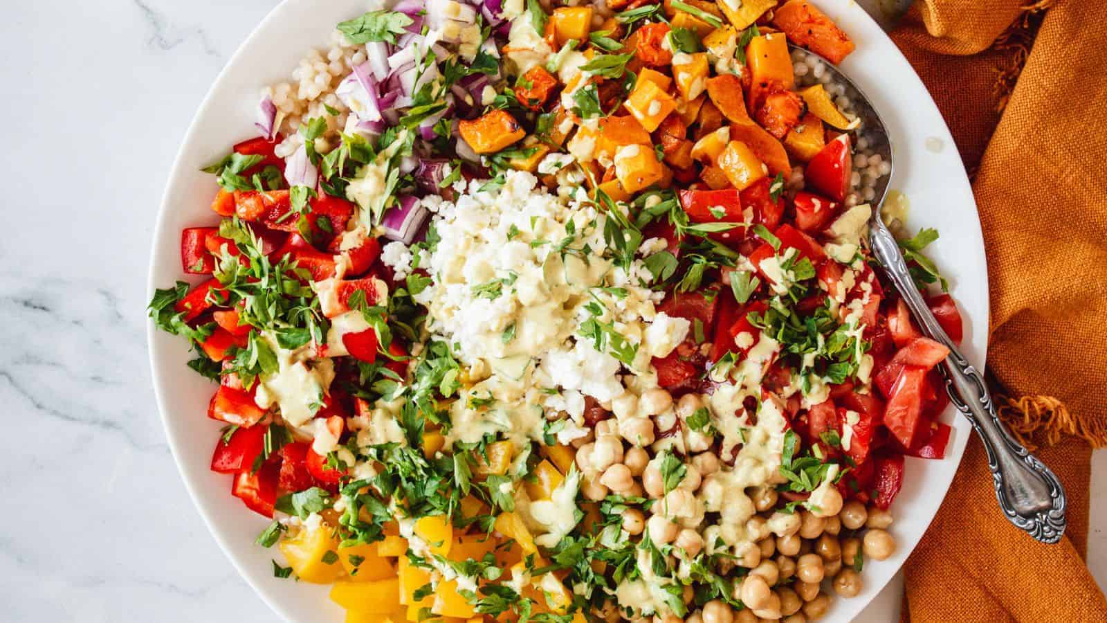 A colorful salad on a white plate featuring chickpeas, diced red and yellow bell peppers, chopped red onions, roasted sweet potatoes, fresh herbs, and a creamy dressing. A spoon is placed on the side. The plate is on a marble surface.