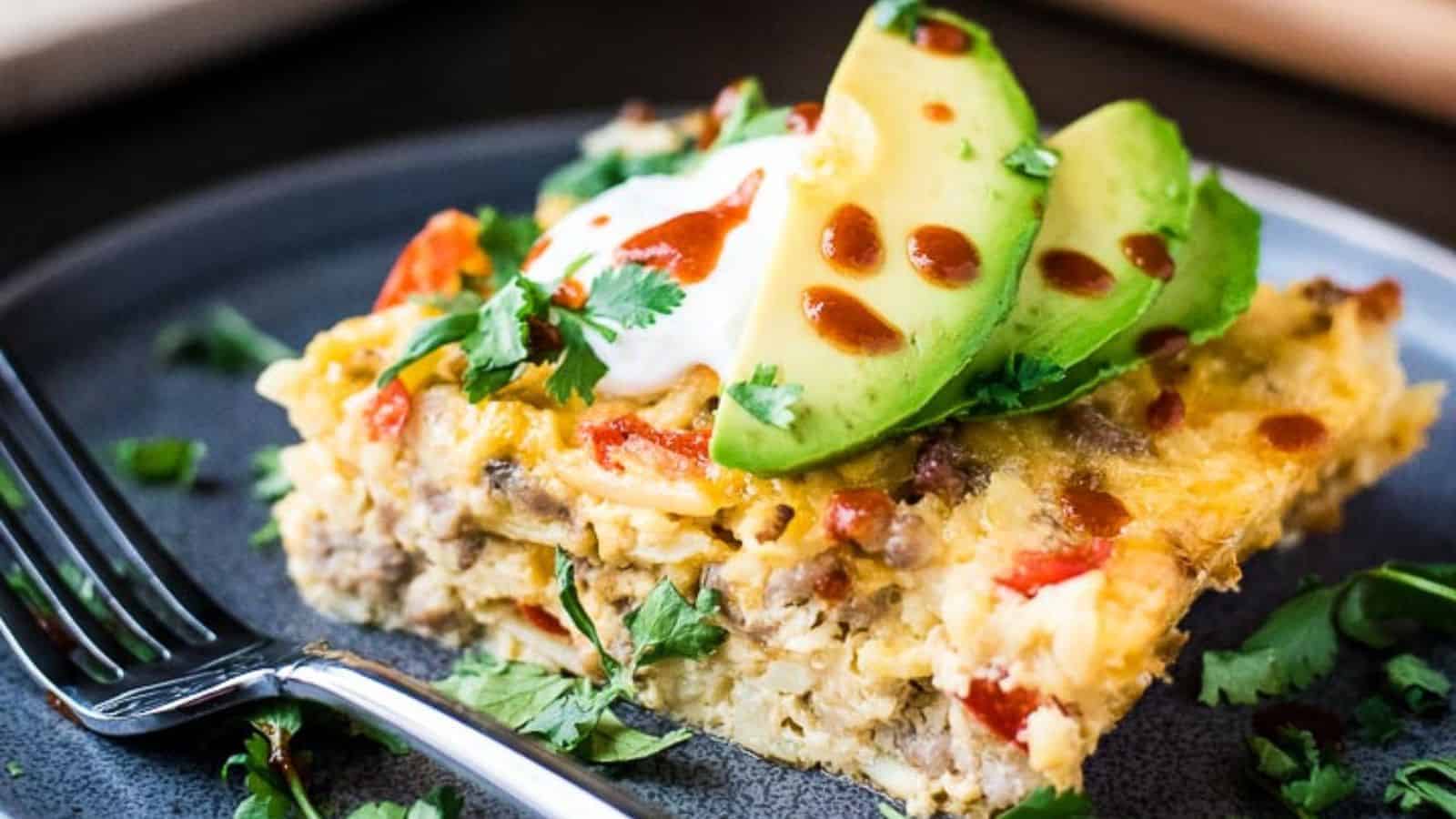 A serving of breakfast casserole on a dark plate, topped with sliced avocado, a dollop of sour cream, and a drizzle of hot sauce. Fresh cilantro is sprinkled on top, with a fork resting beside the savory casserole dish.