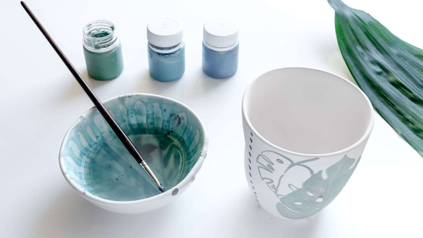 A white bowl with watercolor paint and a brush sits on a table next to a white cup with a painted leaf design. Three small paint jars and a green leaf are in the background on a white surface.