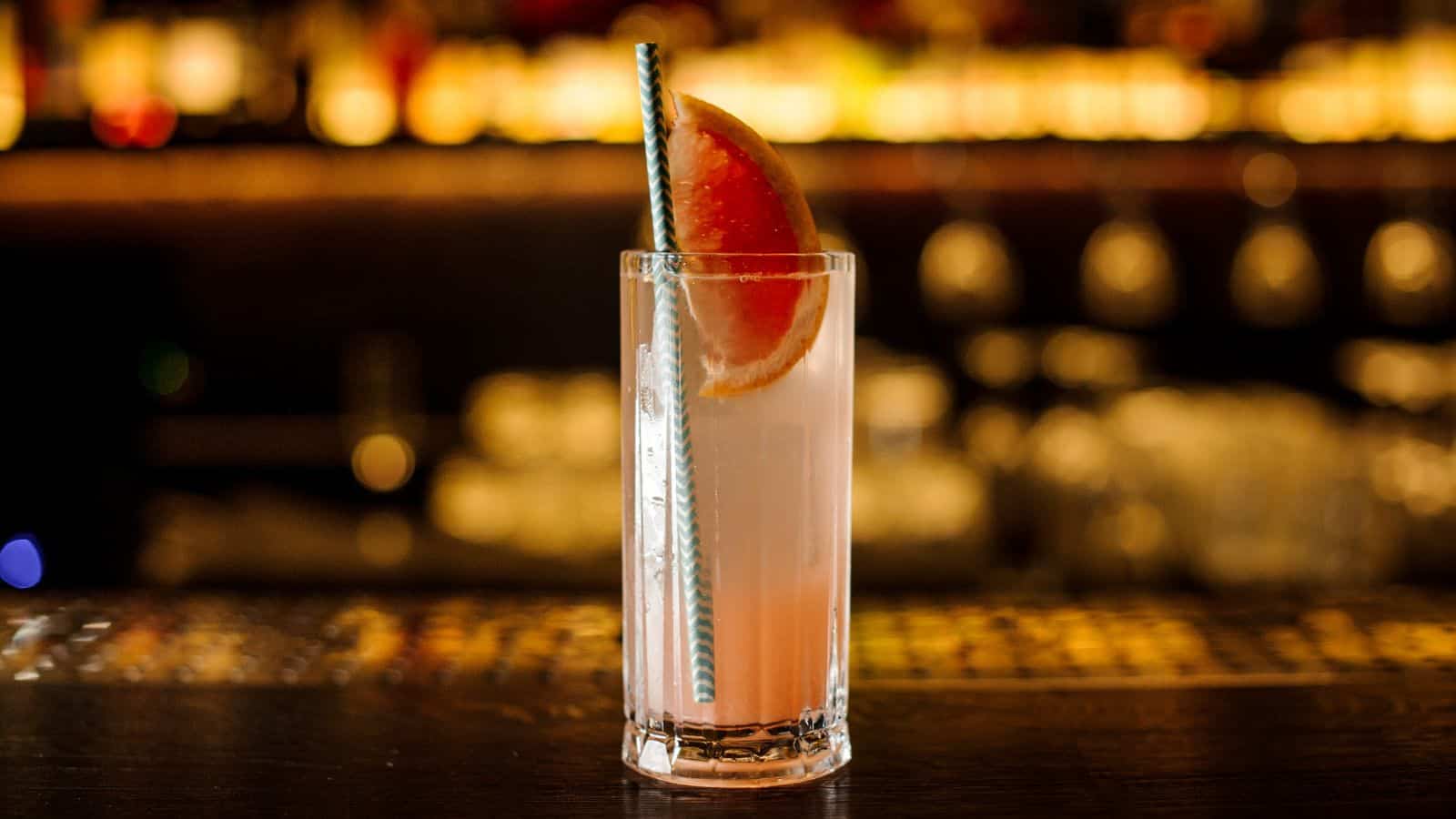 A tall glass filled with a light pink cocktail, garnished with a half slice of grapefruit on the rim and a striped straw. The background is softly blurred, suggesting a bar setting with warm lighting.