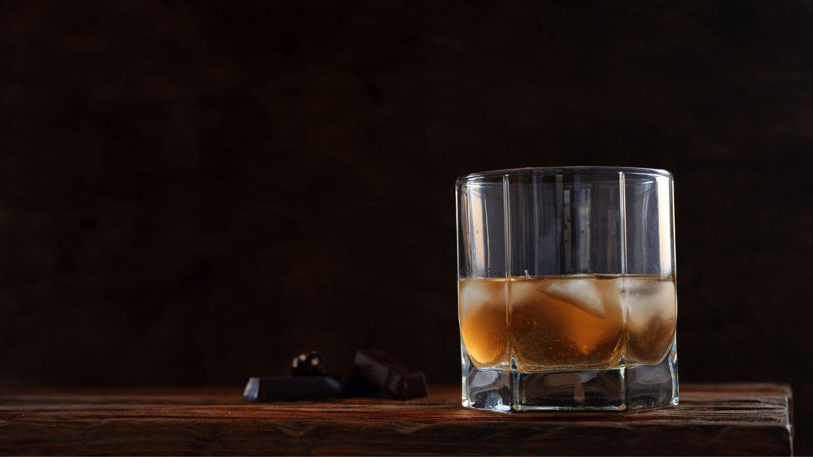 A glass of whiskey with ice cubes sits on a wooden surface. Two pieces of dark chocolate lie nearby. The background is dark and minimal, highlighting the drink and chocolates.