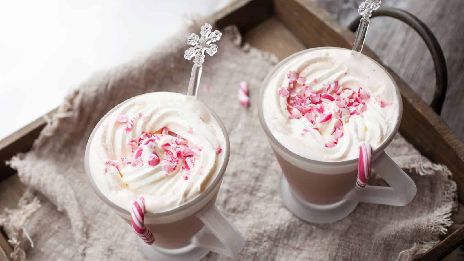 Two glass mugs filled with a creamy beverage topped with whipped cream and pink peppermint pieces. Each mug has a small candy cane on the rim and a decorative stirrer resembling a snowflake, placed on a textured cloth on a wooden tray.