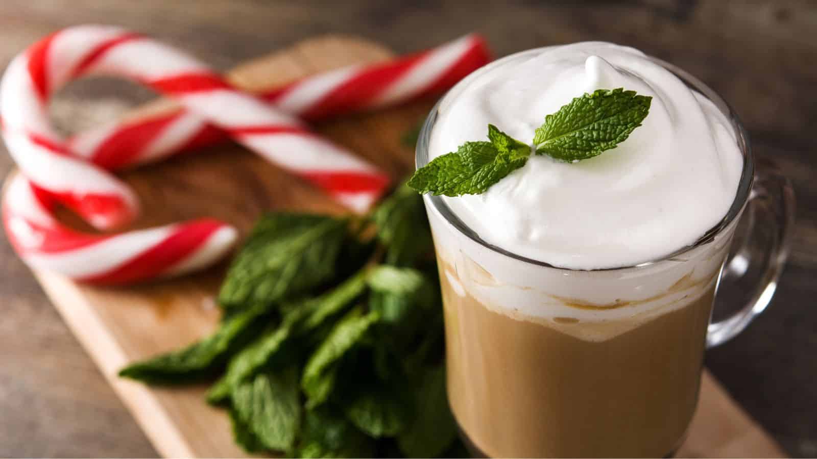 A mug of frothy coffee with a mint garnish sits on a wooden board, surrounded by fresh mint leaves and two striped candy canes.