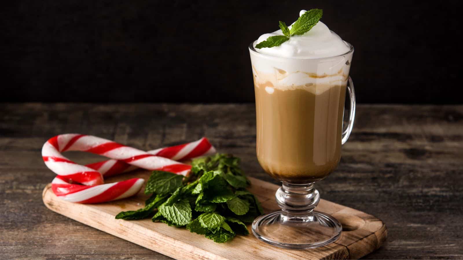 A glass mug filled with a frothy iced coffee sits on a wooden board. It's topped with mint leaves. Next to the mug, there are two candy canes and a bunch of fresh mint leaves. The background is dark and blurred.