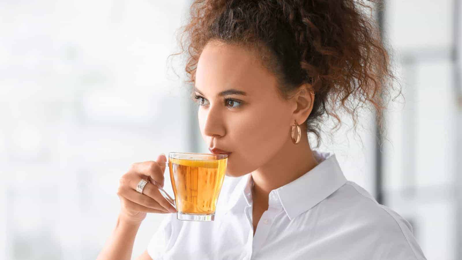 A woman with curly hair, adorned in a white shirt and hoop earrings, sips thoughtfully from a clear glass mug filled with a yellow drink known to increase blood sugar. She gazes to her right in a bright indoor setting, immersed in her surroundings.