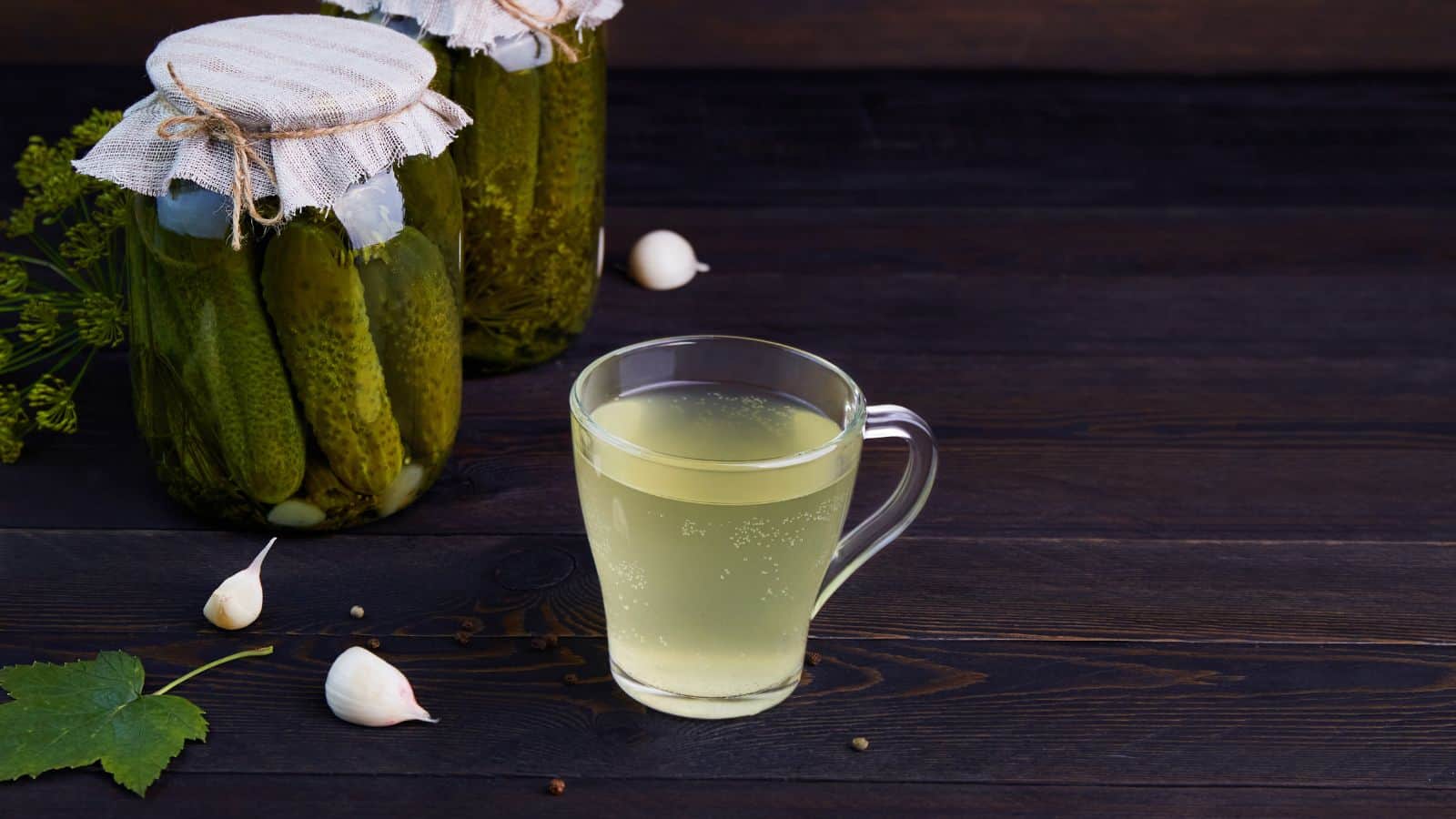 A glass mug filled with a light yellow liquid is placed on a dark wooden surface. Next to it are two jars of pickles covered with fabric and twine. Garlic cloves and a green leaf are scattered around on the table.