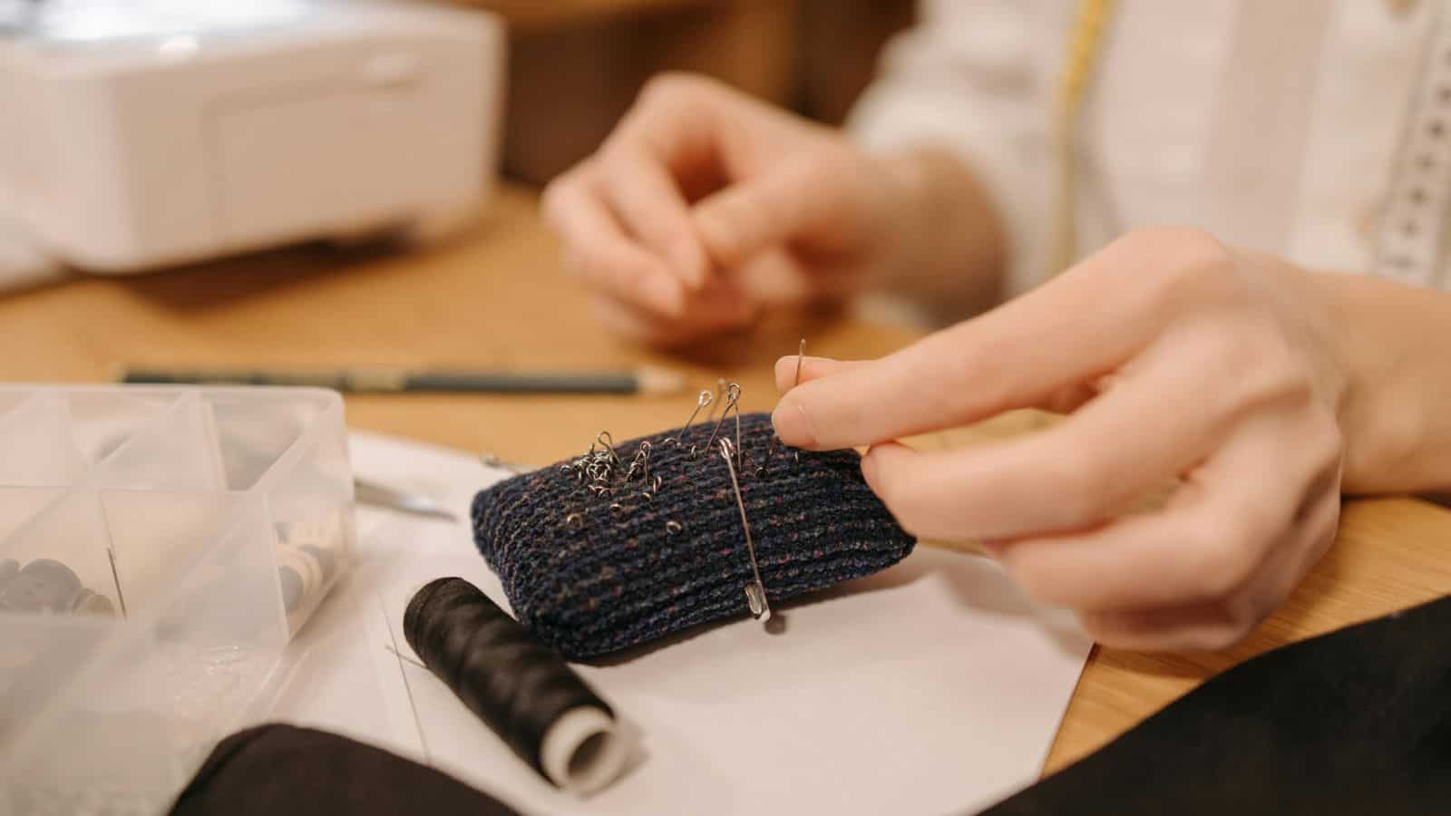 A person's hands are sewing, using a needle and pin cushion. A spool of black thread and a sewing machine are on the wooden table. A storage box with sewing supplies is partially visible.