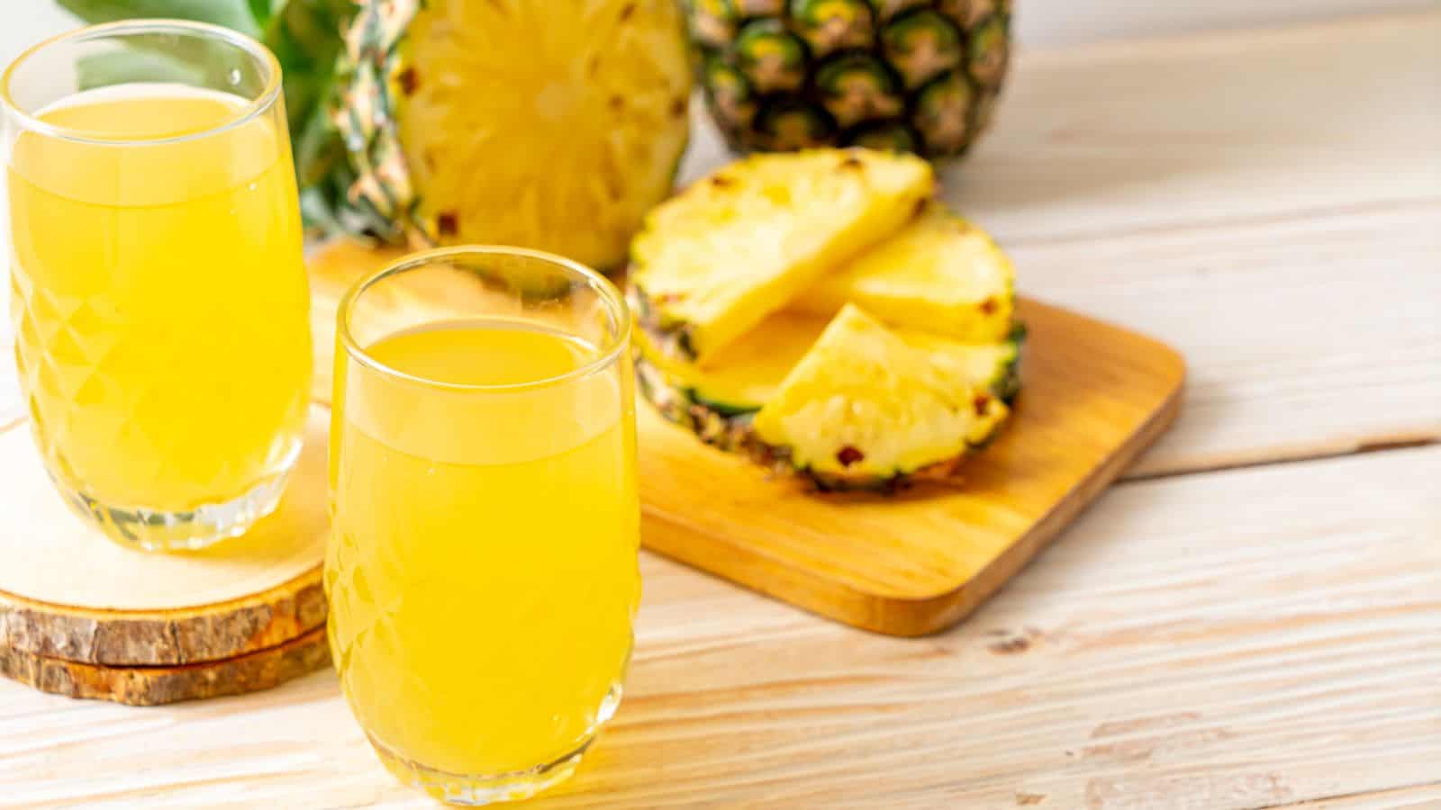 Two glasses of pineapple juice sit on a wooden surface next to a cutting board with sliced pineapple. A whole pineapple is partially visible in the background. The setting is bright and fresh, suggesting a summery theme.