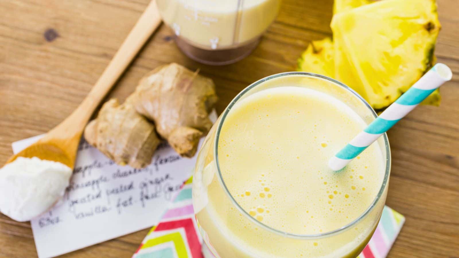 A glass of yellow smoothie with a blue and white striped straw sits on a colorful napkin. Fresh ginger, pineapple slices, a wooden spoon with yogurt, and a handwritten recipe are in the background on a wooden surface.