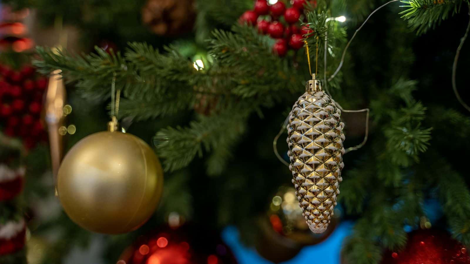 A decorated Christmas tree featuring a gold bauble and a pine cone-shaped ornament. Red berries and smaller red ornaments are visible in the background, with green branches filling the scene.