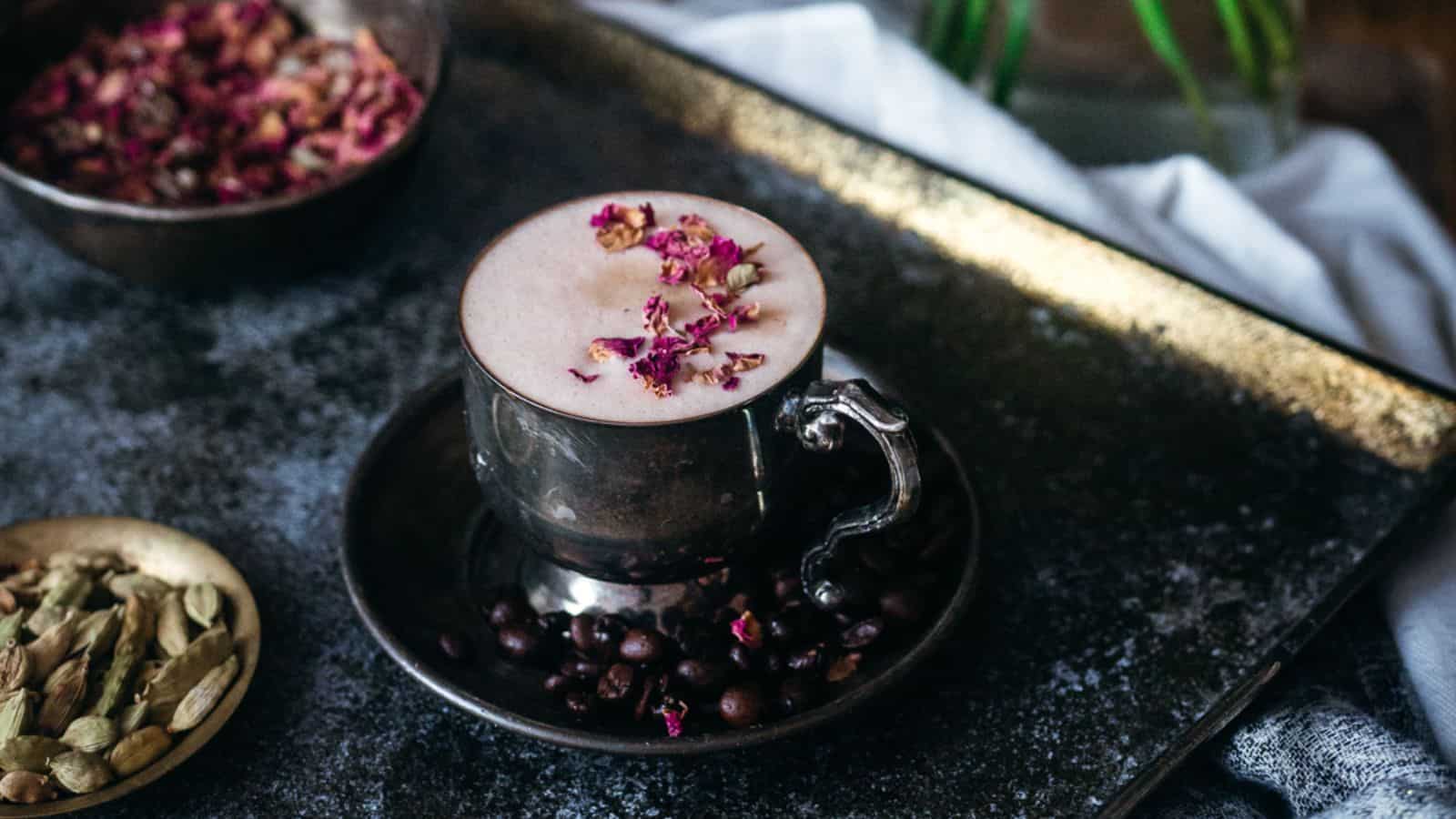 A metal cup of frothy coffee or tea is on a dark tray, garnished with rose petals. Coffee beans are scattered around the cup. Nearby are a bowl of dried rose petals and a plate with cardamom pods.