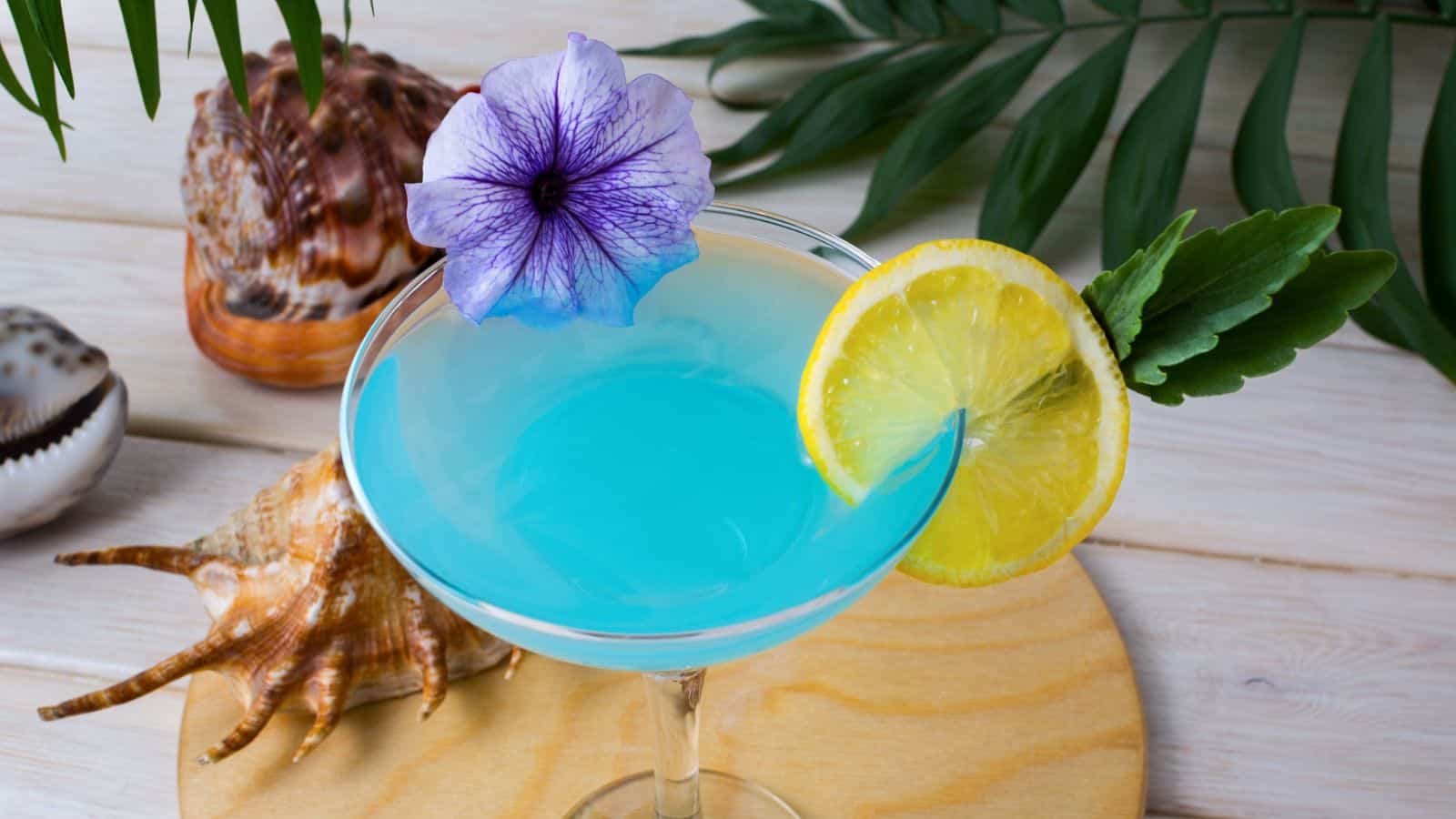 A blue cocktail in a martini glass garnished with a slice of lemon, a purple flower, and a green leaf. The glass is placed on a wooden surface with seashells and tropical leaves as decoration.