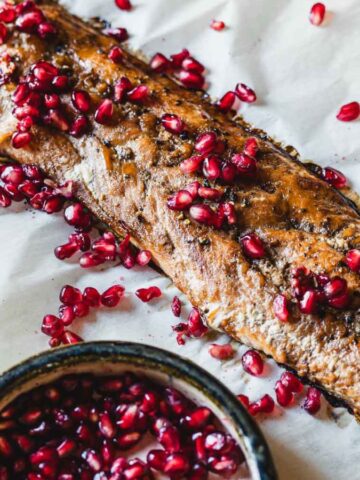 A baked fish fillet on a parchment-lined baking sheet is garnished with scattered pomegranate seeds. A small bowl of additional pomegranate seeds is placed nearby.