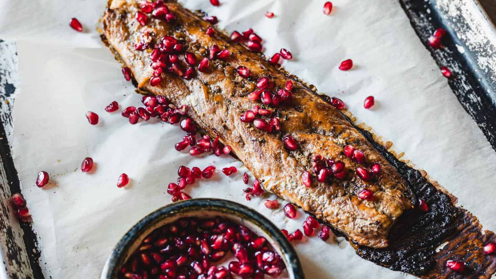 A baked fish fillet on a parchment-lined baking sheet is garnished with scattered pomegranate seeds. A small bowl of additional pomegranate seeds is placed nearby.