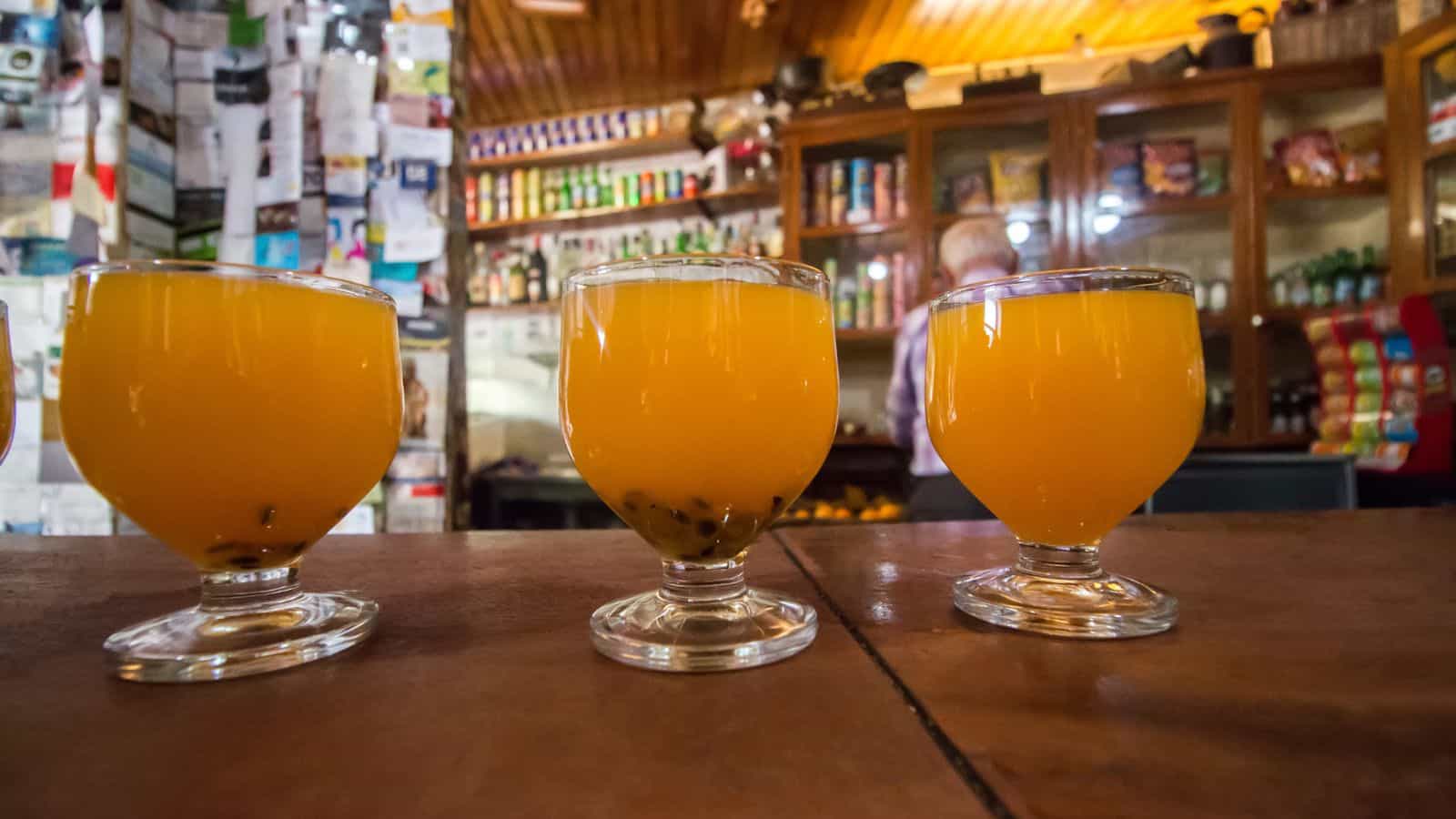 Three glasses of passion fruit juice with seeds at the bottom are placed on a wooden counter. Shelves stocked with various items and a person in the background are visible in a cozy, warmly lit room.