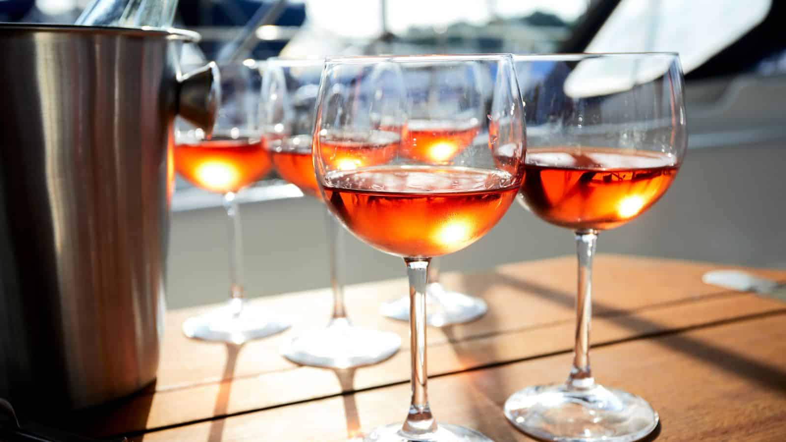 Several glasses of ros&eacute; wine are placed on a wooden table, with sunlight reflecting off the liquid. A metal ice bucket is partially visible on the left side. The background is slightly blurred, conveying a relaxed, outdoor atmosphere.