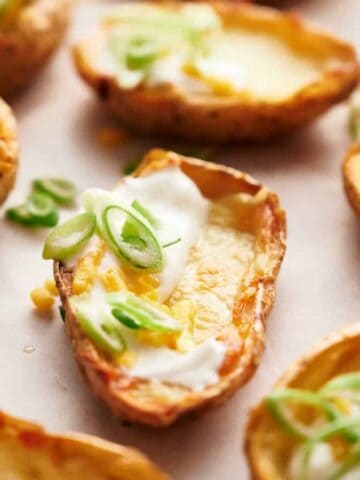 Potato skins on parchment paper, topped with melted cheese, sour cream, and slices of green onion.
