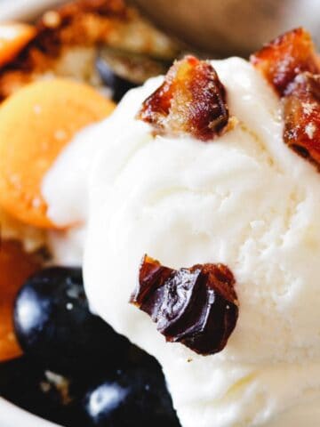 A bowl of dessert features a scoop of vanilla ice cream topped with chopped dates. Surrounding the ice cream are fresh golden and dark purple grapes. A spoon is placed in the bowl, resting on a blue surface.