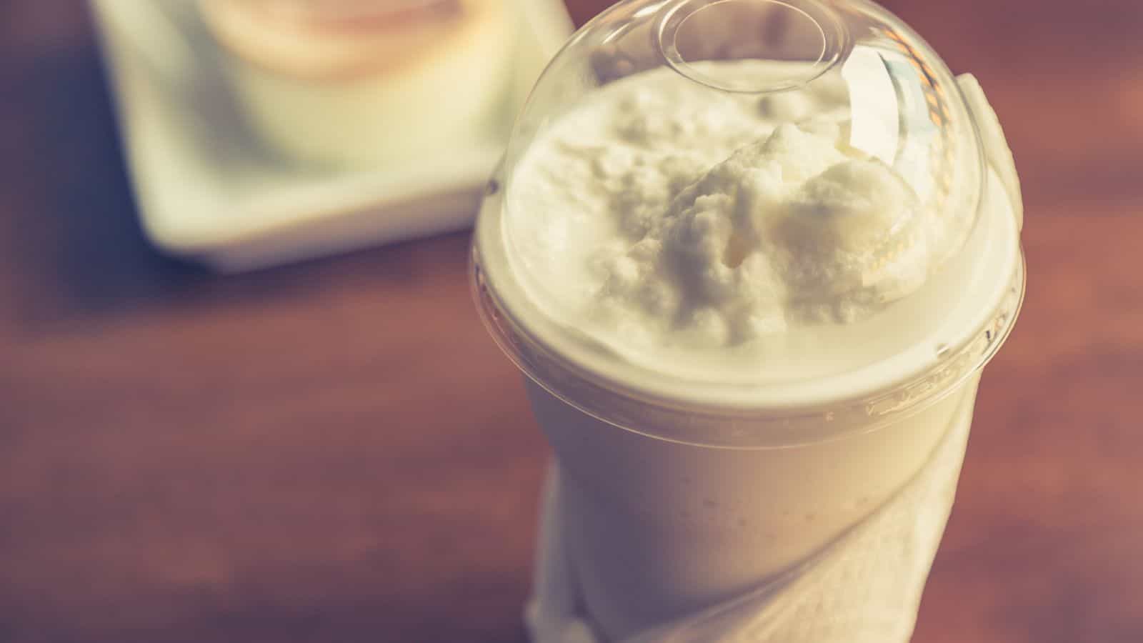 A close-up of a plastic cup with a dome lid containing a creamy white beverage, likely a milkshake or smoothie. The cup is wrapped in a napkin. In the blurred background, there is a small dish with a dessert on a wooden table.