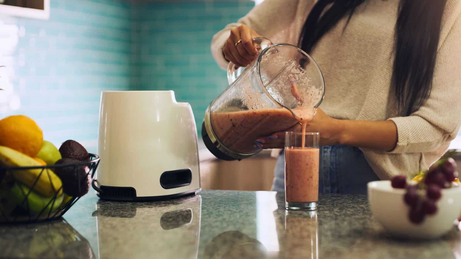A person pours a pink smoothie from a blender into a glass on a kitchen counter. The blender base is nearby. A fruit basket containing oranges, avocados, and other fruits is in the background. The kitchen has a tiled backsplash.