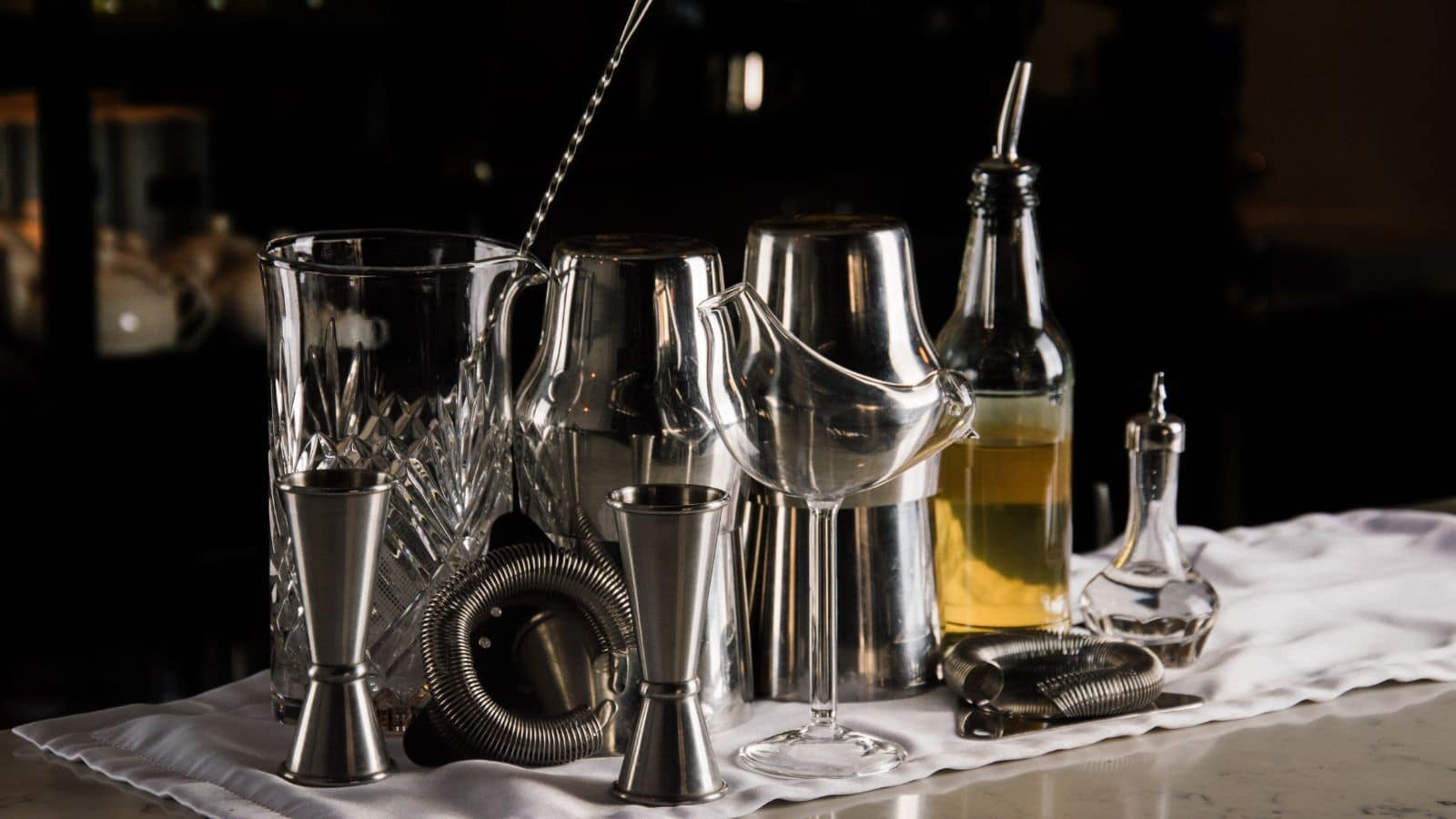 A collection of bar tools is arranged on a white cloth. It includes shakers, a mixing glass, jiggers, a strainer, a tall stirring spoon, a bottle with liquid, and a coupe glass. The background is dark, highlighting the shiny metal surfaces.