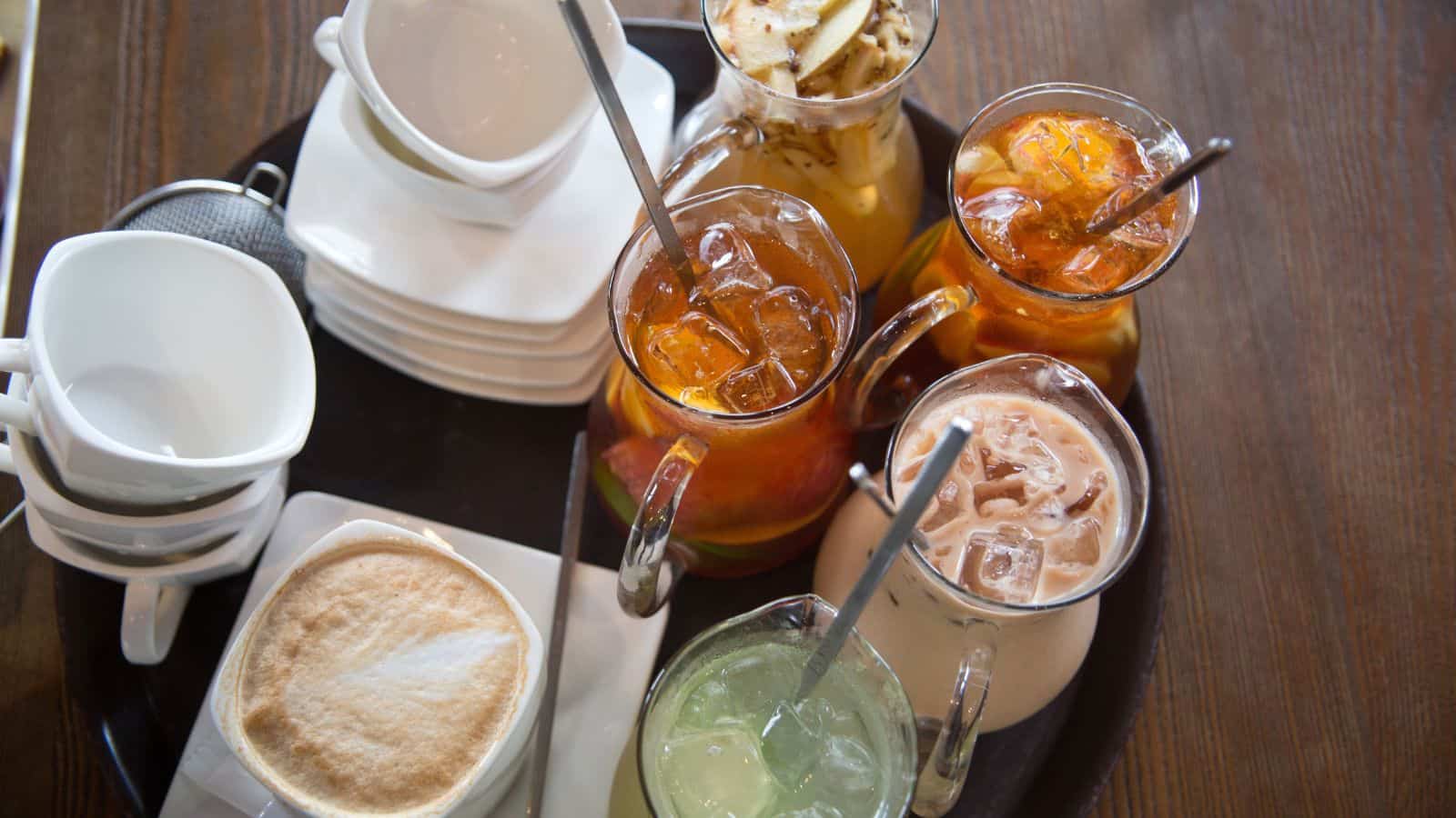 A tray containing various beverages, including iced tea with fruit slices, iced coffee, a cappuccino, a light green drink, and empty stacked cups with saucers, placed on a wooden table.