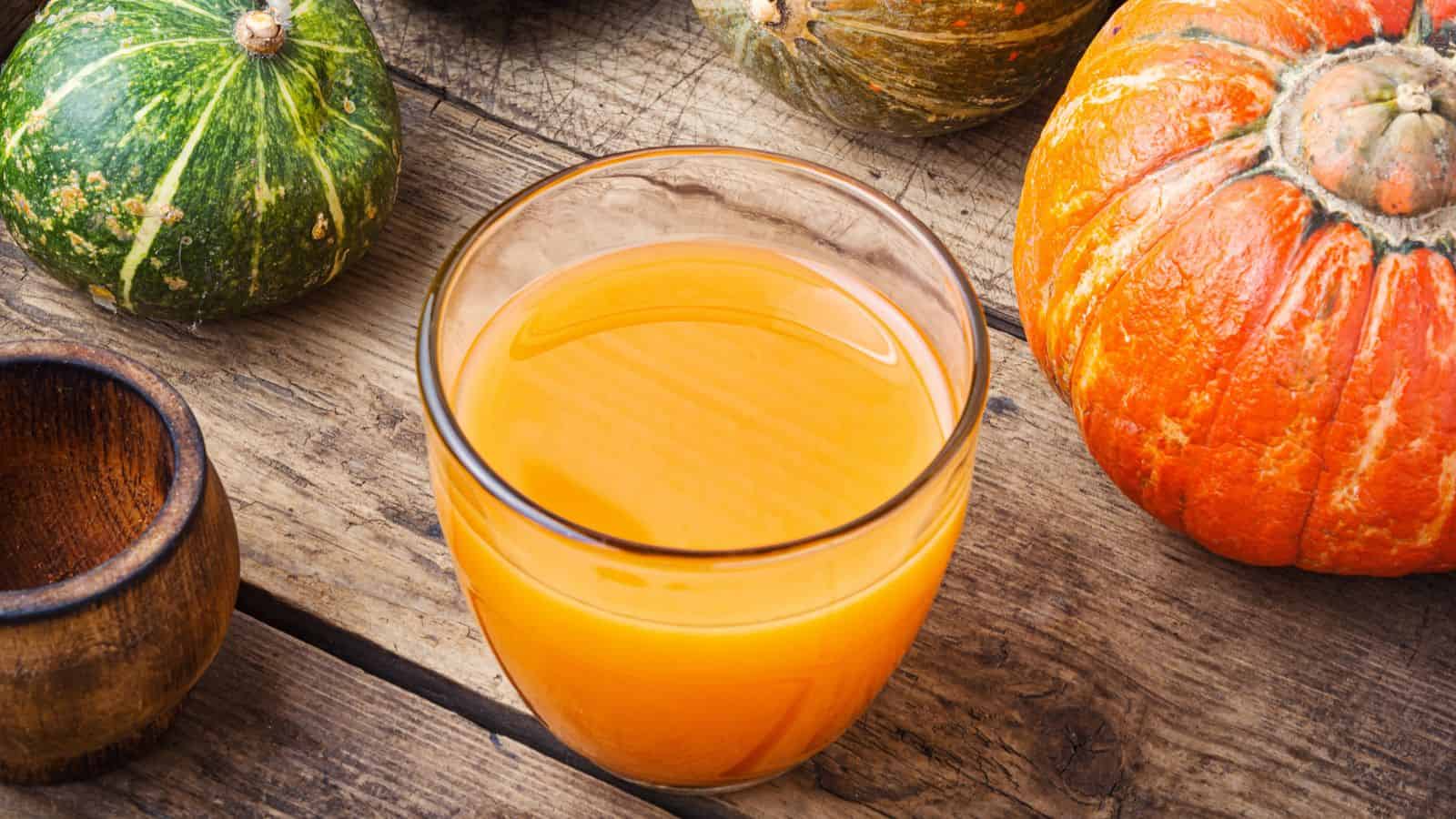 A glass of orange pumpkin juice on a wooden surface, surrounded by various whole pumpkins in green and orange hues. A small wooden bowl is partially visible on the left. The scene gives an autumnal, rustic feel.