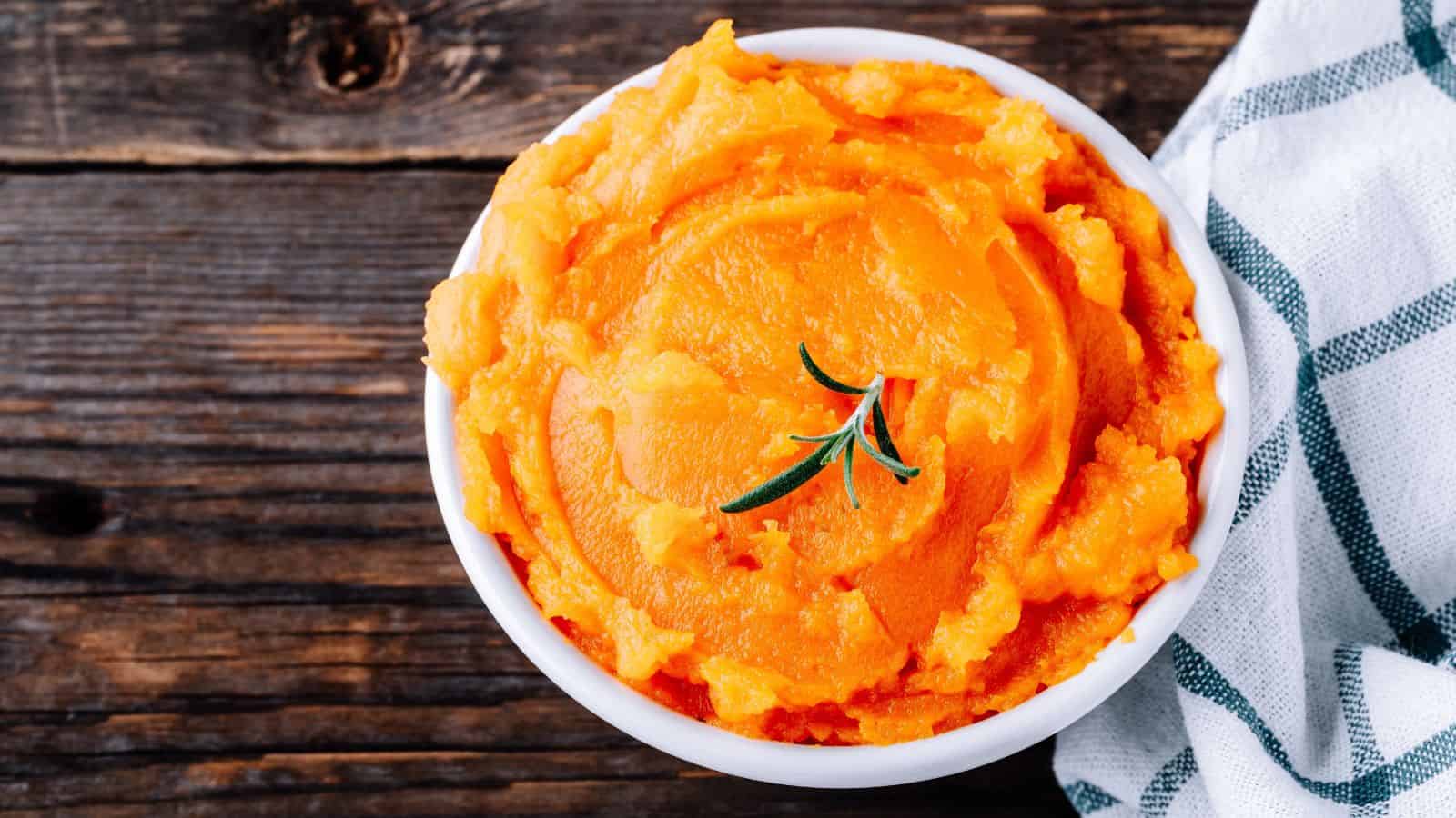 A bowl of mashed sweet potatoes is garnished with a sprig of rosemary. It is placed on a wooden surface next to a white and grey cloth.