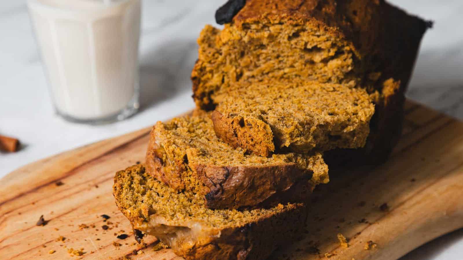 Sliced pumpkin bread on a wooden cutting board next to a glass of milk.