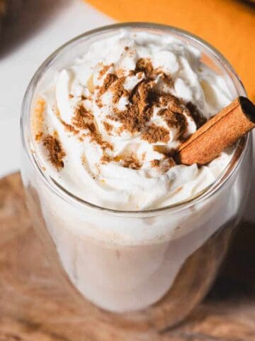 A glass mug of whipped cream-topped beverage sprinkled with cinnamon and garnished with a cinnamon stick sits on a wooden tray. A pumpkin and an orange cloth are in the blurred background.