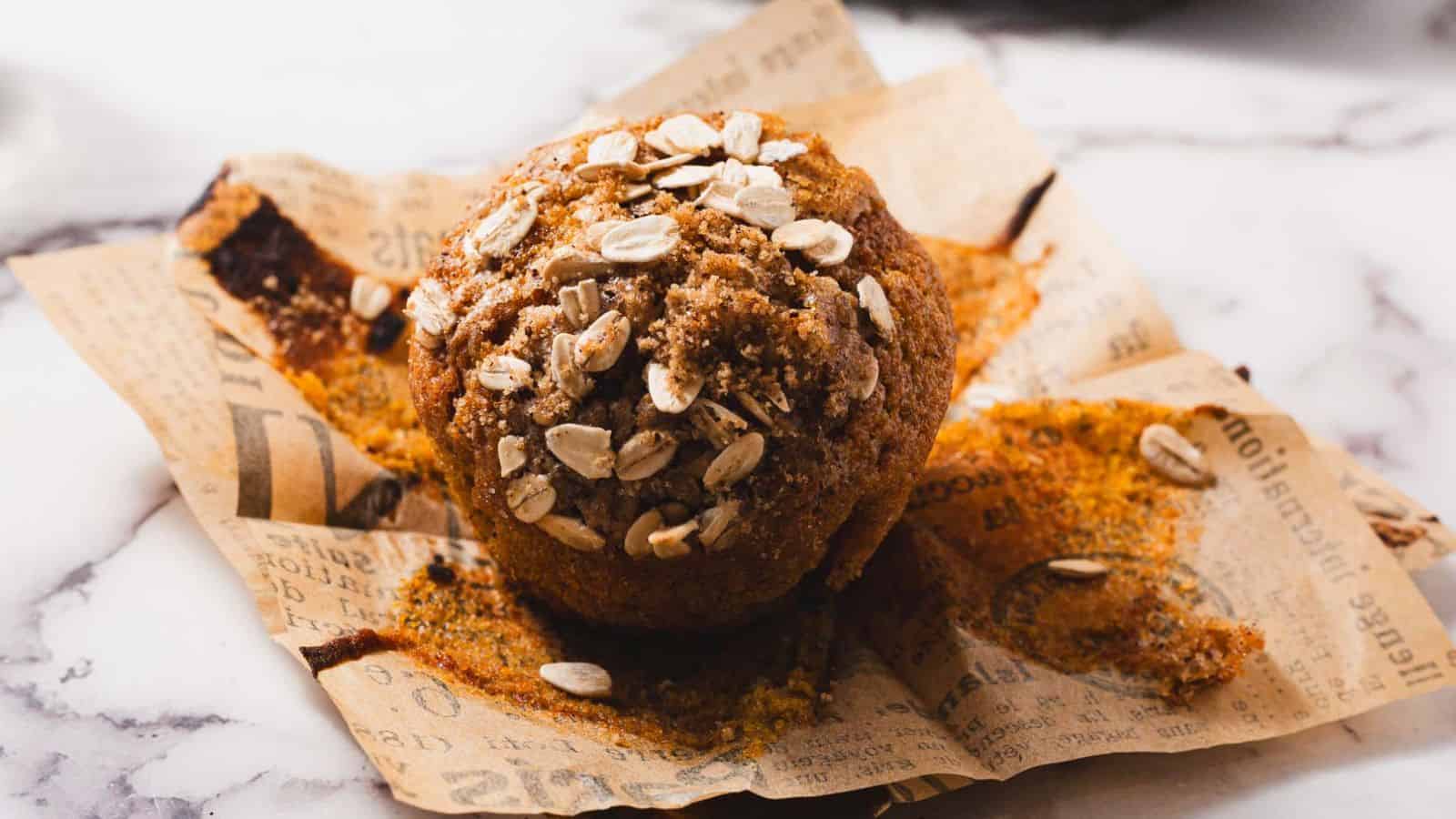 A close-up of a muffin topped with oats, resting on crumpled brown paper with printed text. The muffin is set on a marble surface.