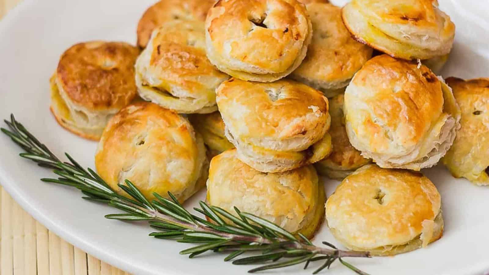 A plate of golden-brown puff pastries stacked on a white dish, garnished with a sprig of rosemary.