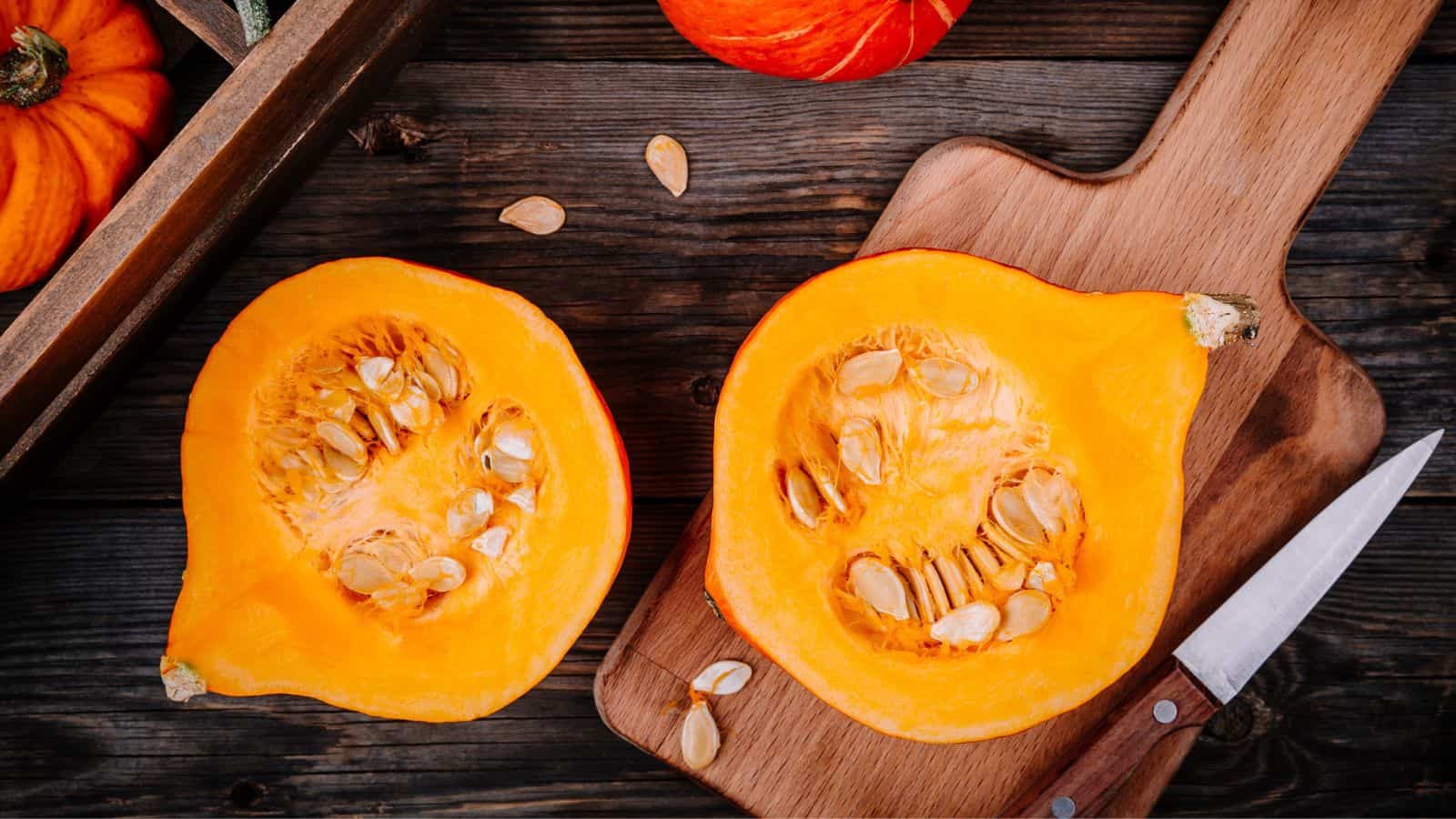 A halved pumpkin showing its seeds and inner texture is placed on a wooden cutting board. A knife lies nearby. Additional pumpkins and seeds are scattered on the wooden surface in the background.