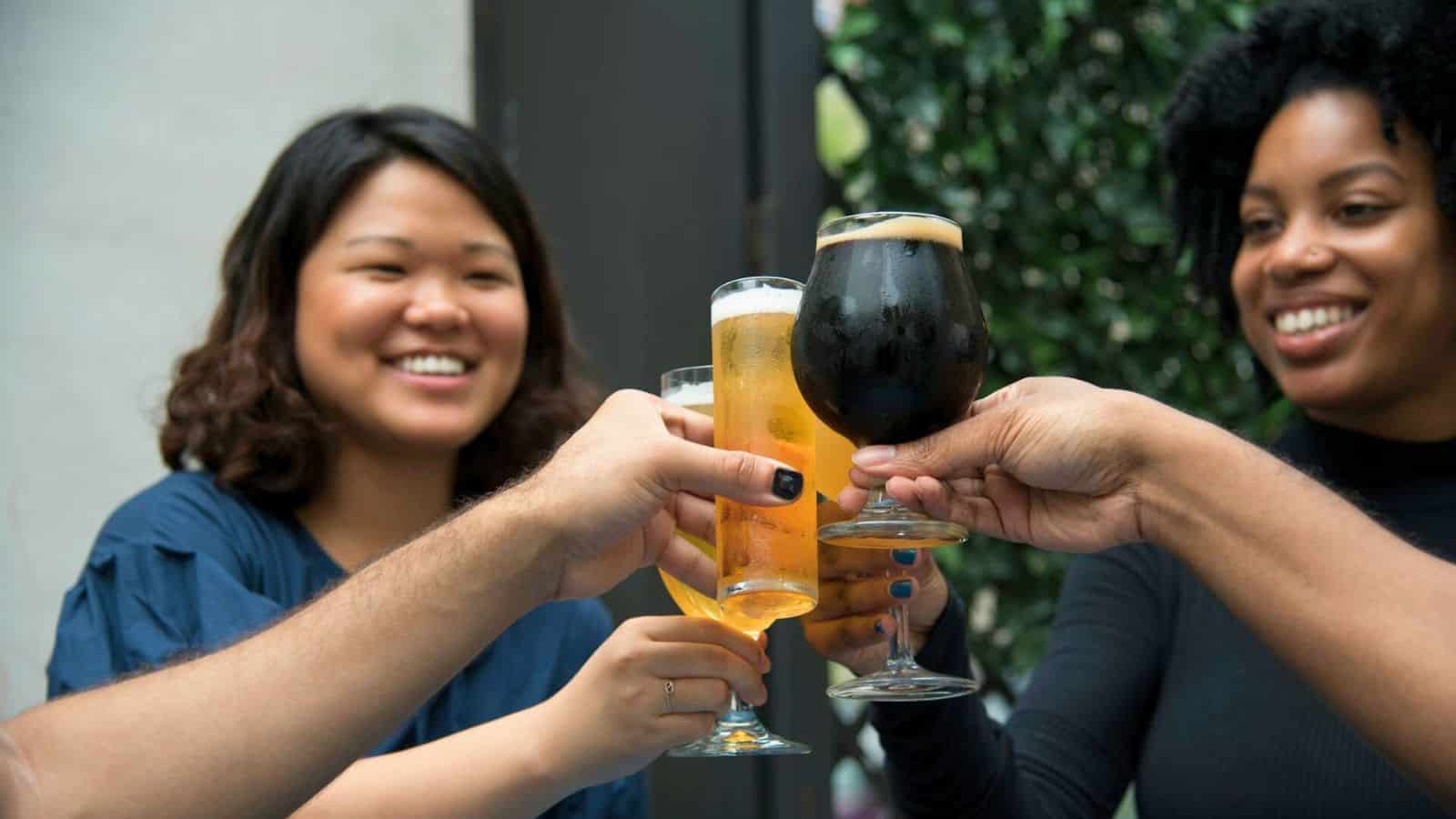 Three people clink glasses while smiling in a casual setting. They are holding different types of beverages, including beer and a dark stout. Greenery is visible in the background.