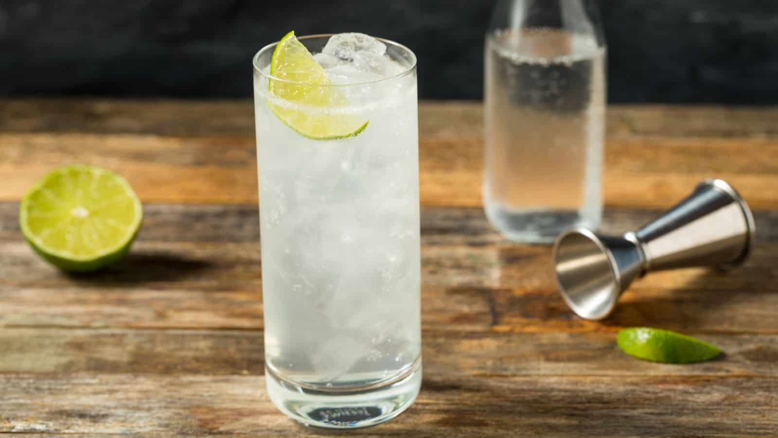 A tall glass filled with a clear sparkling drink and ice, garnished with a lime wedge, sits on a wooden surface. Nearby are a lime half, a metal jigger, and a clear bottle.