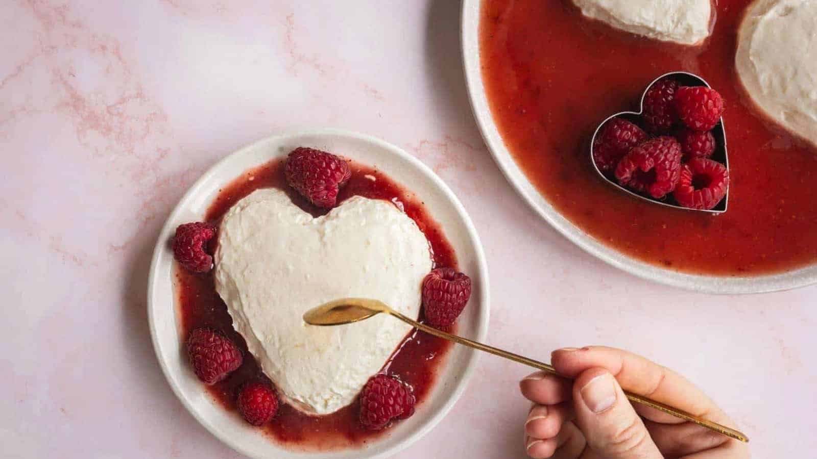 A heart-shaped dessert topped with cream and surrounded by raspberries on a white plate. Next to it is a larger plate with similar desserts and a metal mold filled with raspberries. A hand holds a small spoon over the dessert.