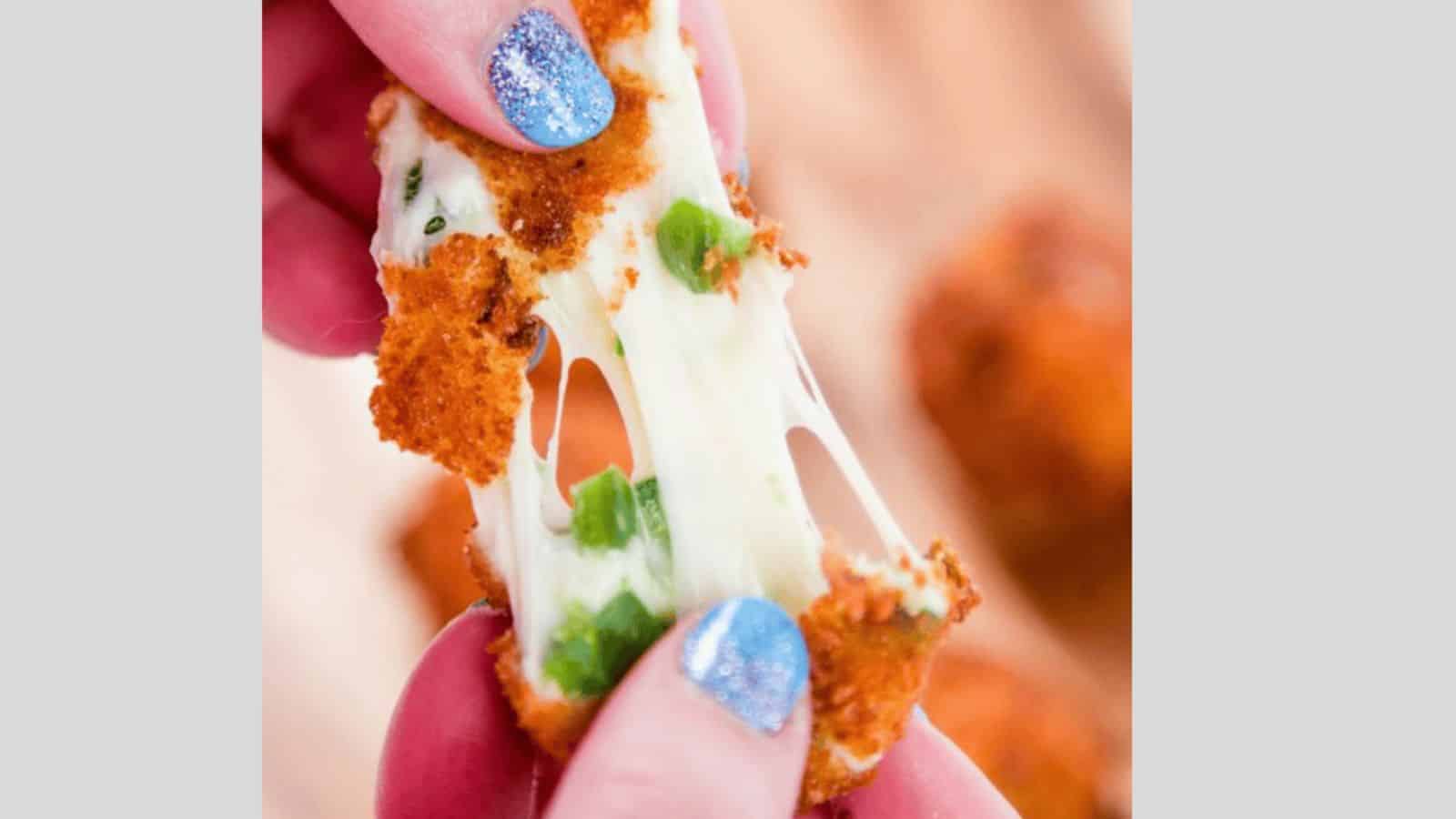 A close-up of a person pulling apart a breaded and fried cheese stick. Melted cheese is stretching between the two pieces, with some green herbs visible. The person has blue glitter nail polish.