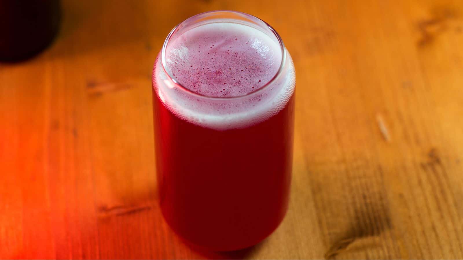 A glass filled with frothy red juice sits on a wooden surface. The beverage has a bubbly foam on top, and the wooden table has a warm, reddish hue due to the lighting.
