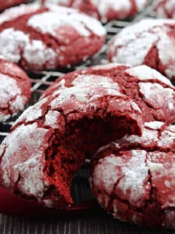 Red velvet crinkle cookies dusted with powdered sugar are arranged on a cooling rack. One cookie in the foreground has a bite taken out. A red ribbon is woven around the cookies, creating a decorative look.
