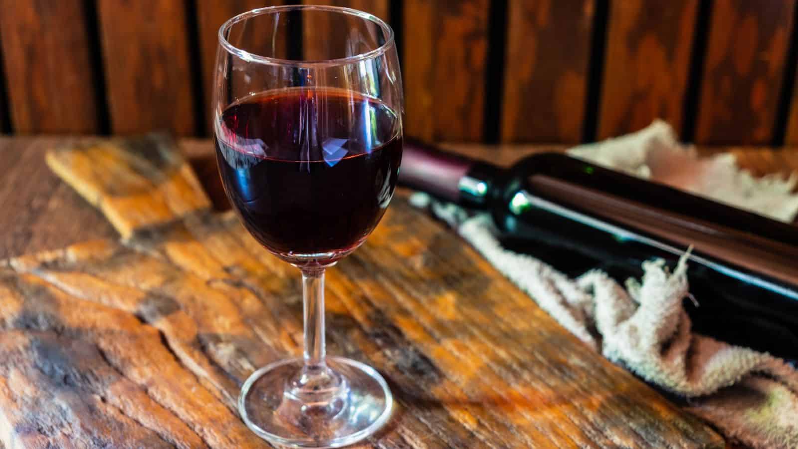 A glass of red wine is placed on a wooden surface beside a wine bottle wrapped in a cloth. The setting includes a rustic wooden background.