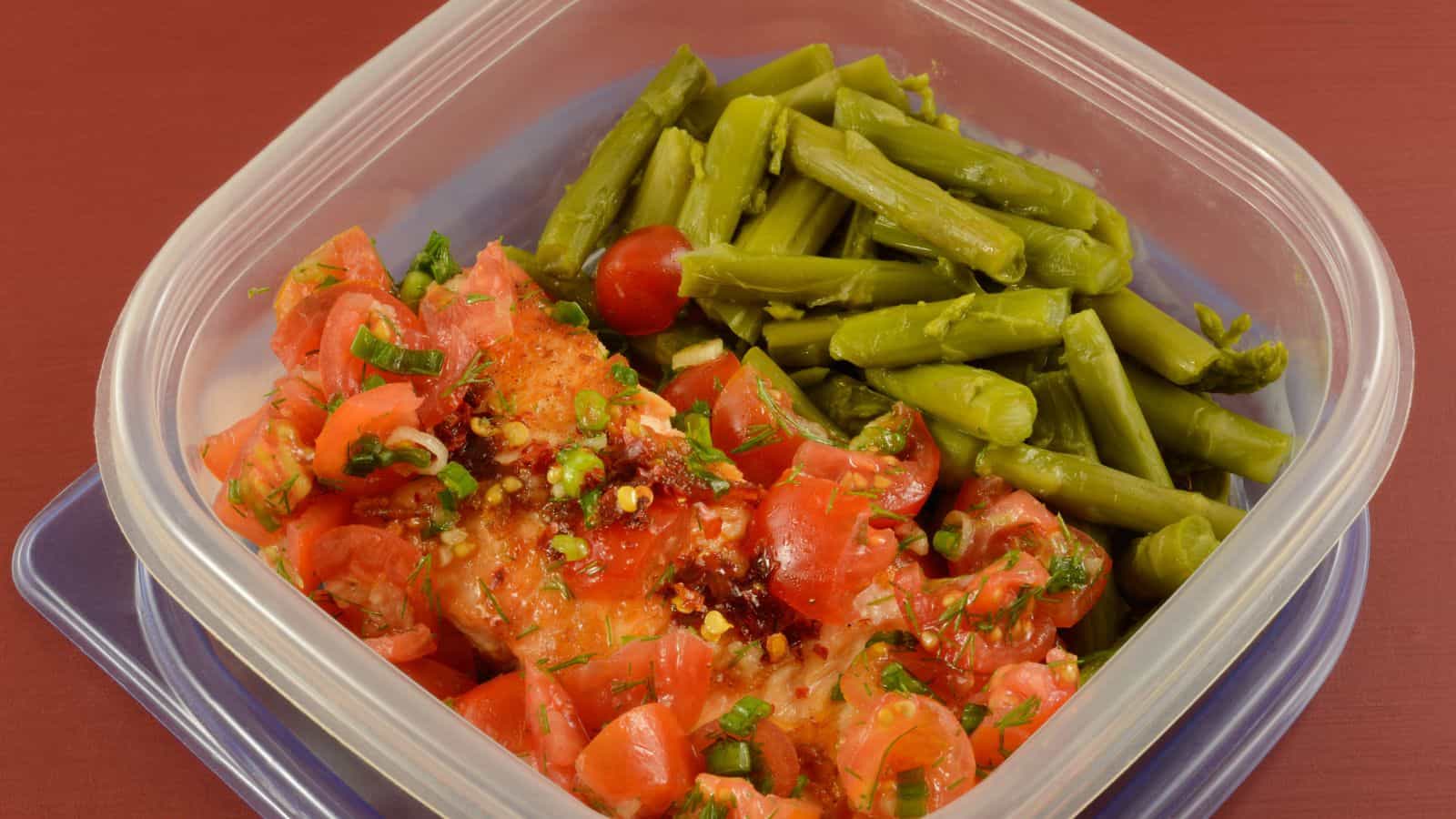 A clear plastic container with cooked green asparagus spears on one side and diced tomatoes mixed with herbs on the other side, set against a red background.