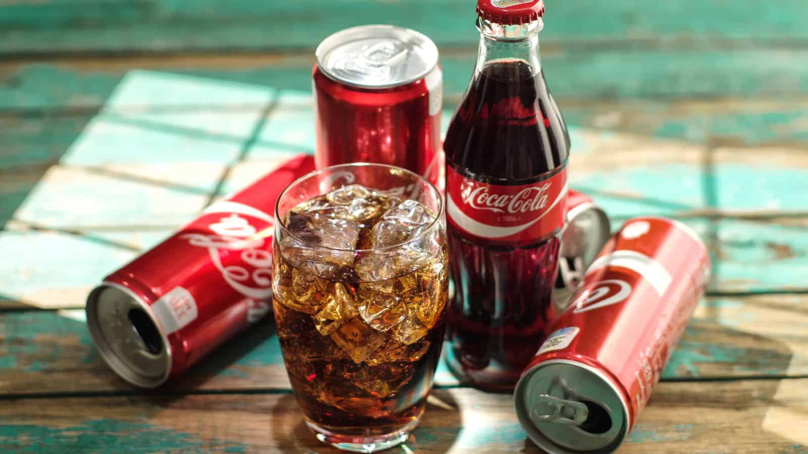 A glass filled with ice and Coca-Cola is in the foreground. Behind it, there is a Coca-Cola bottle and three red cans. One can is open, and two are closed, all on a wooden surface with a turquoise hue.