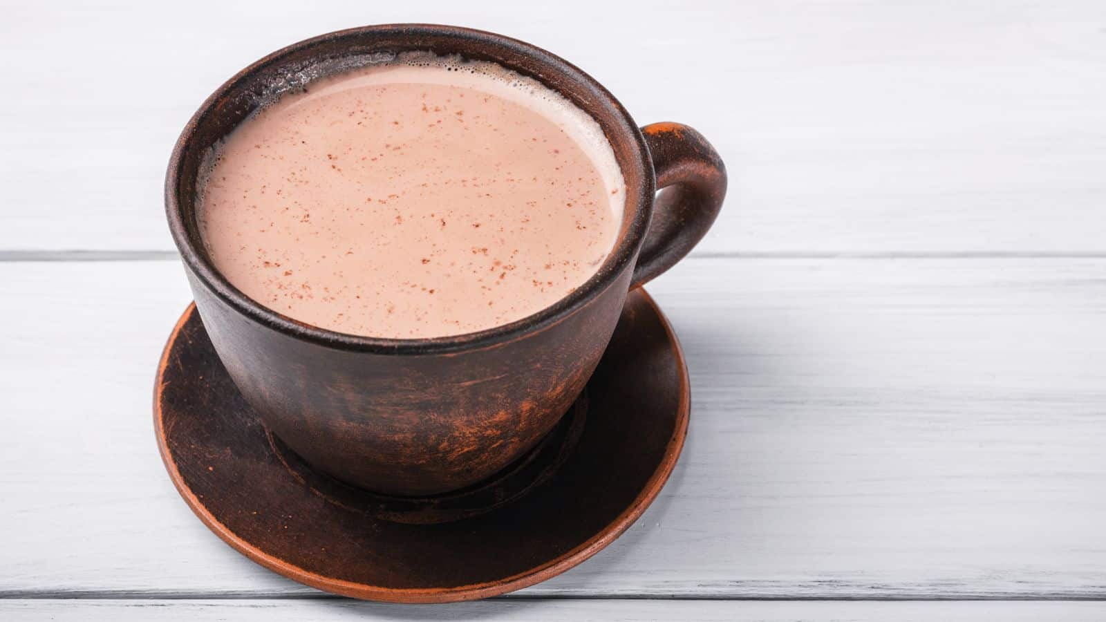 A brown ceramic cup filled with foamy hot chocolate is placed on a matching saucer. The setup rests on a white wooden surface.
