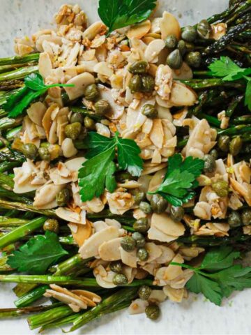 A plate of roasted asparagus topped with sliced almonds, capers, and fresh parsley. Two lemon wedges are placed on the side of the oval plate.