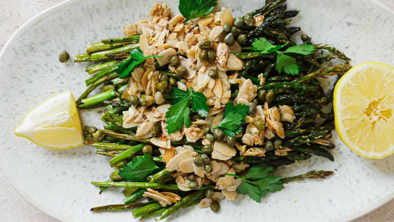 A plate of roasted asparagus topped with sliced almonds, capers, and fresh parsley. Two lemon wedges are placed on the side of the oval plate.
