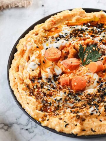 A plate of hummus garnished with slices of cherry tomato, a parsley leaf, and sprinkled with black sesame seeds and spices. The dish is placed on a marble surface next to a partial view of a plate with sliced bread.