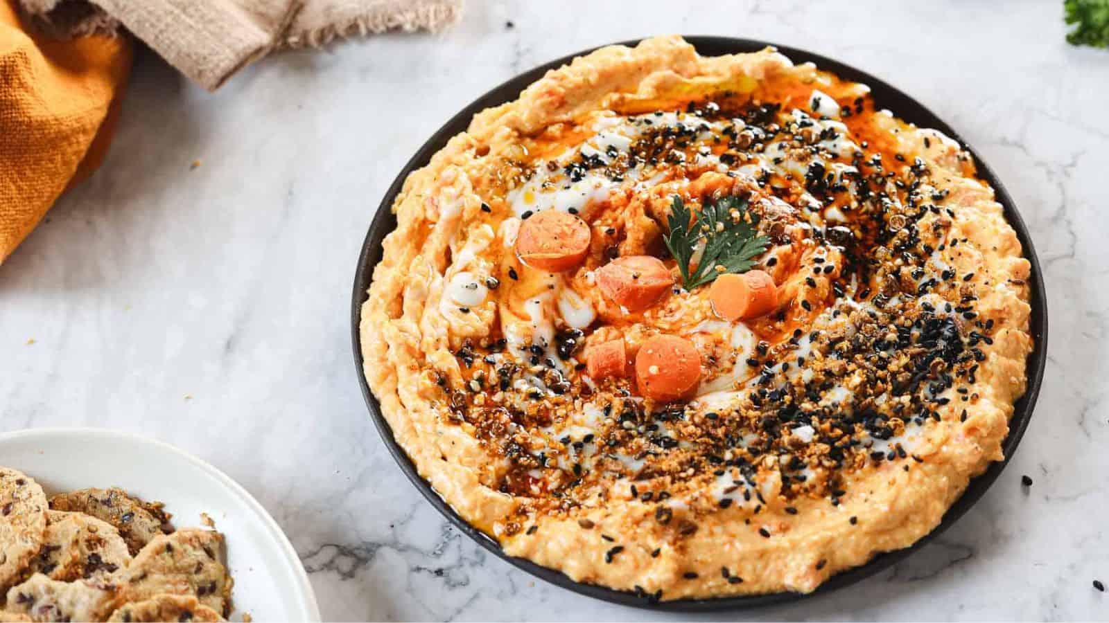 A plate of hummus garnished with slices of cherry tomato, a parsley leaf, and sprinkled with black sesame seeds and spices. The dish is placed on a marble surface next to a partial view of a plate with sliced bread.