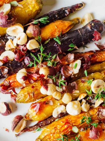 A plate of roasted multicolored carrots topped with chopped hazelnuts and garnished with fresh thyme. The carrots are glazed and appear slightly caramelized, arranged neatly on a white dish.