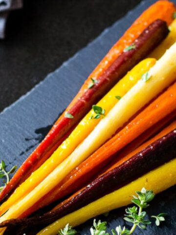 A slate serving board with a bunch of roasted heirloom carrots in shades of orange, purple, and yellow. Fresh herbs are scattered on top, and a sprig of thyme lies beside them. A fork rests in the background.