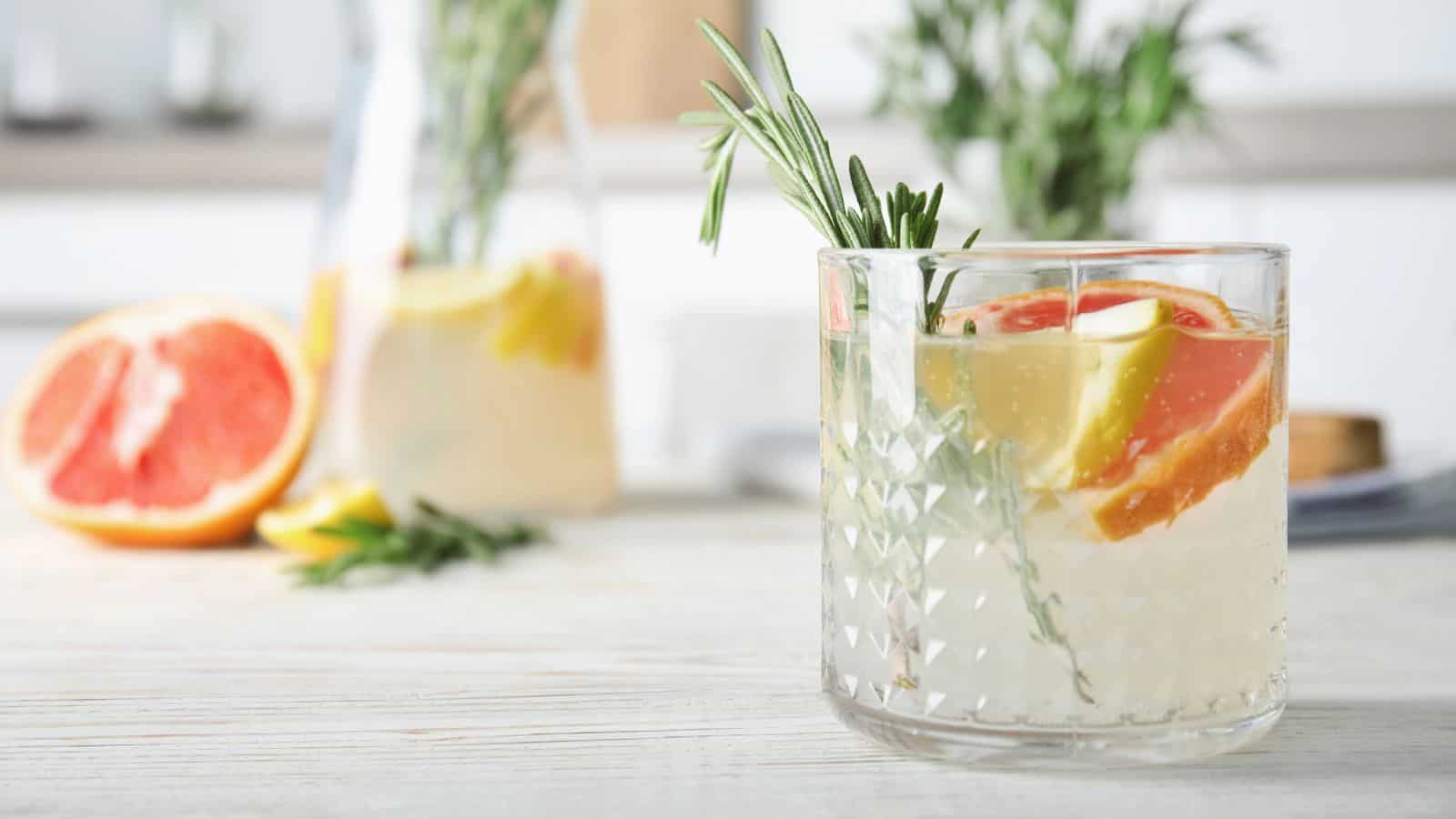 A glass of infused water with slices of grapefruit and lemon is garnished with rosemary. In the background, a pitcher with more infused water and sliced fruits is visible. A halved grapefruit and rosemary sprigs are on the table.